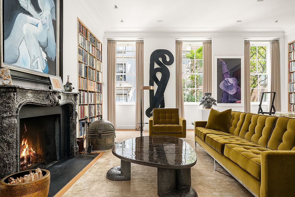 This sophisticated living room features a prominent stone fireplace with an ornate mantel, flanked by built-in bookshelves that span the wall. A plush, olive-toned velvet sofa and matching armchair provide comfortable seating around a unique, dark-toned sculptural coffee table. Large windows with neutral drapery allow natural light to fill the space, highlighting the curated art pieces and the elegant, textured rug.
