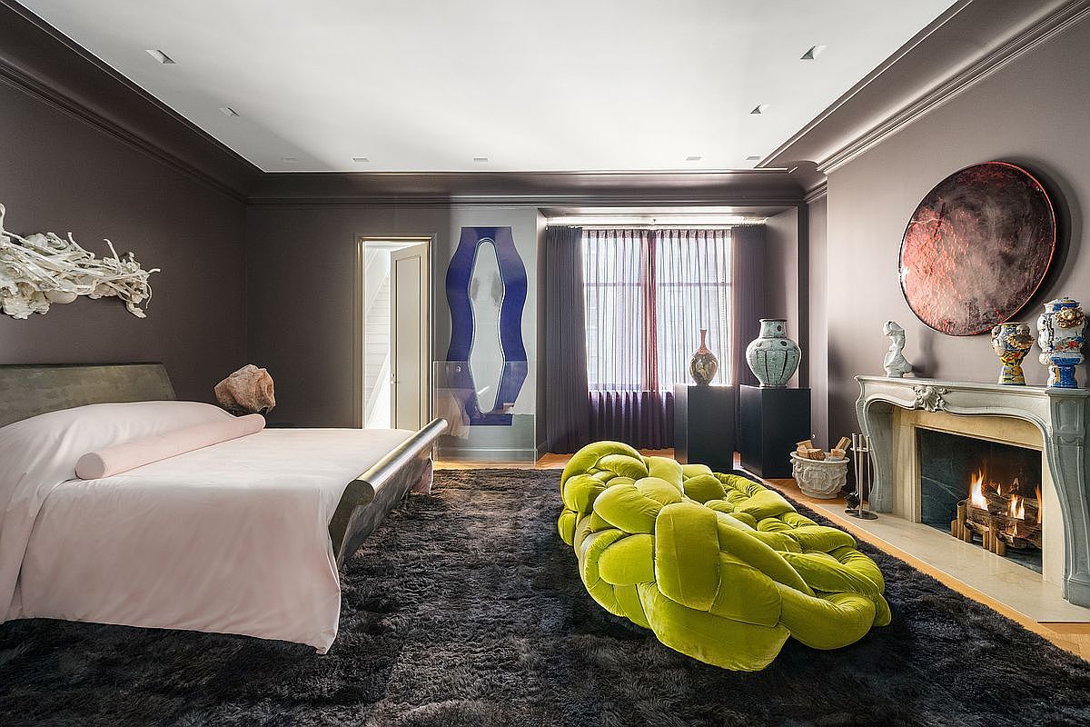 This sophisticated primary bedroom features dark, moody walls and a plush, dark-toned area rug that anchors the space. A prominent lime-green, knotted-style sculptural seating piece sits in the center of the room, contrasting with the soft pink bedding on the bed. The room is accented by a traditional fireplace with a decorative mantel, a large circular piece of wall art, and a unique blue, wave-shaped mirror near the window.