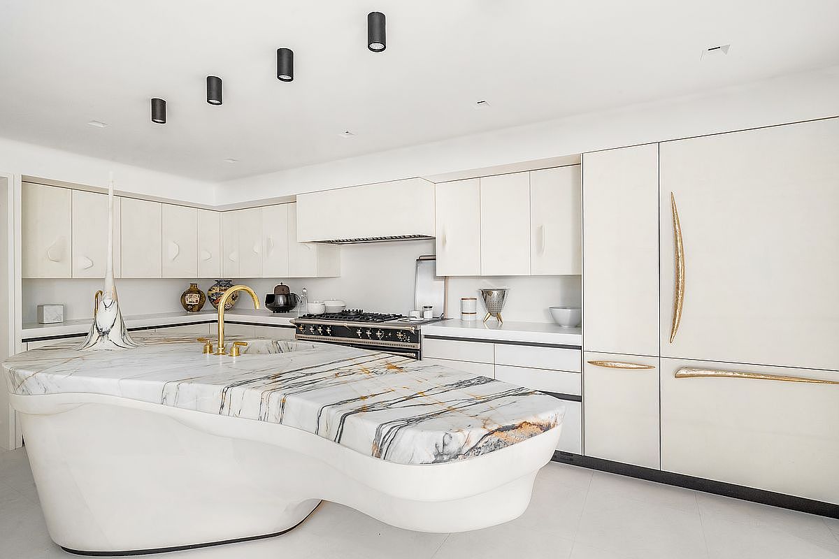 This ultra-modern kitchen features a striking, sculptural island with a dramatic, veined stone countertop that curves into a white base. The space is defined by sleek, handleless white cabinetry, a professional-grade range, and gold-toned hardware accents. Recessed ceiling lights provide a clean, minimalist aesthetic, creating a bright and sophisticated atmosphere.