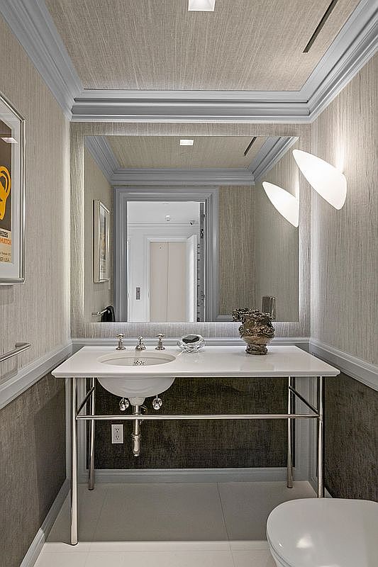 This elegant powder room features a sleek, pedestal-style vanity with a white countertop and polished metal legs. The walls and ceiling are finished in a textured, neutral-toned wallcovering, complemented by a large, framed mirror and modern, sculptural wall sconces. The space is designed with a sophisticated, minimalist aesthetic, emphasizing clean lines and high-end finishes.