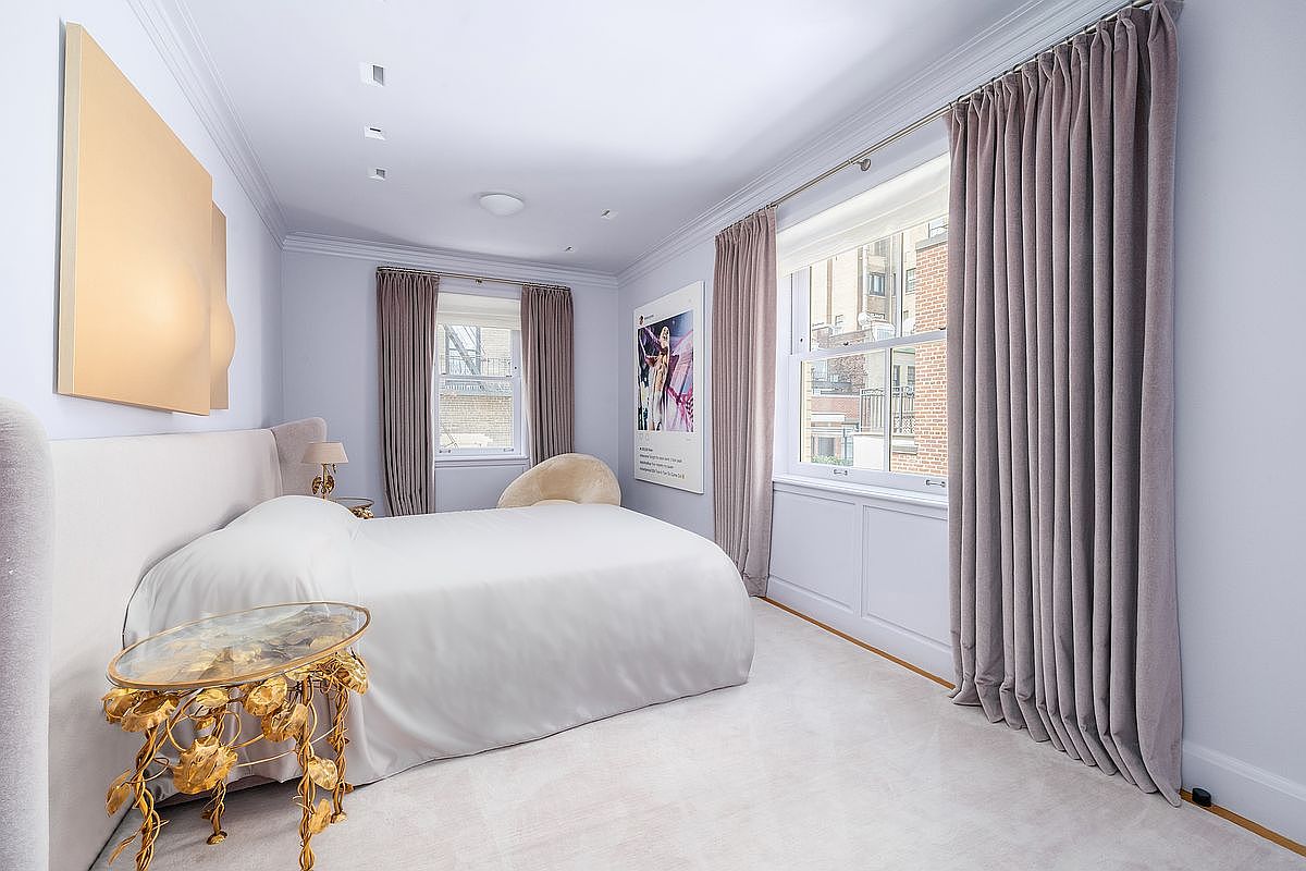 This elegant primary bedroom features a neutral color palette with soft lavender-toned walls and plush carpeting. The room is anchored by a large bed with a light-colored upholstered headboard, complemented by a unique gold-toned side table with a glass top. Large windows draped in floor-length curtains allow for natural light, while framed artwork adds a personalized touch to the sophisticated space.