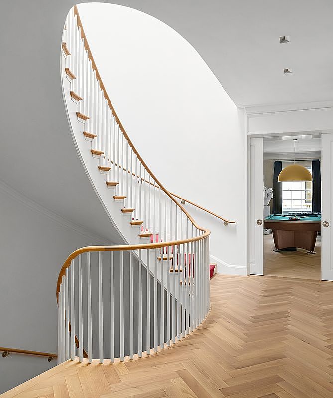 This elegant interior space features a striking curved staircase with white balusters and a light wood handrail, set against a smooth, minimalist white wall. The flooring is composed of light wood planks arranged in a classic herringbone pattern, leading toward a doorway that reveals a game room with a pool table. The perspective is from the landing, emphasizing the architectural flow and the bright, airy atmosphere of the home.