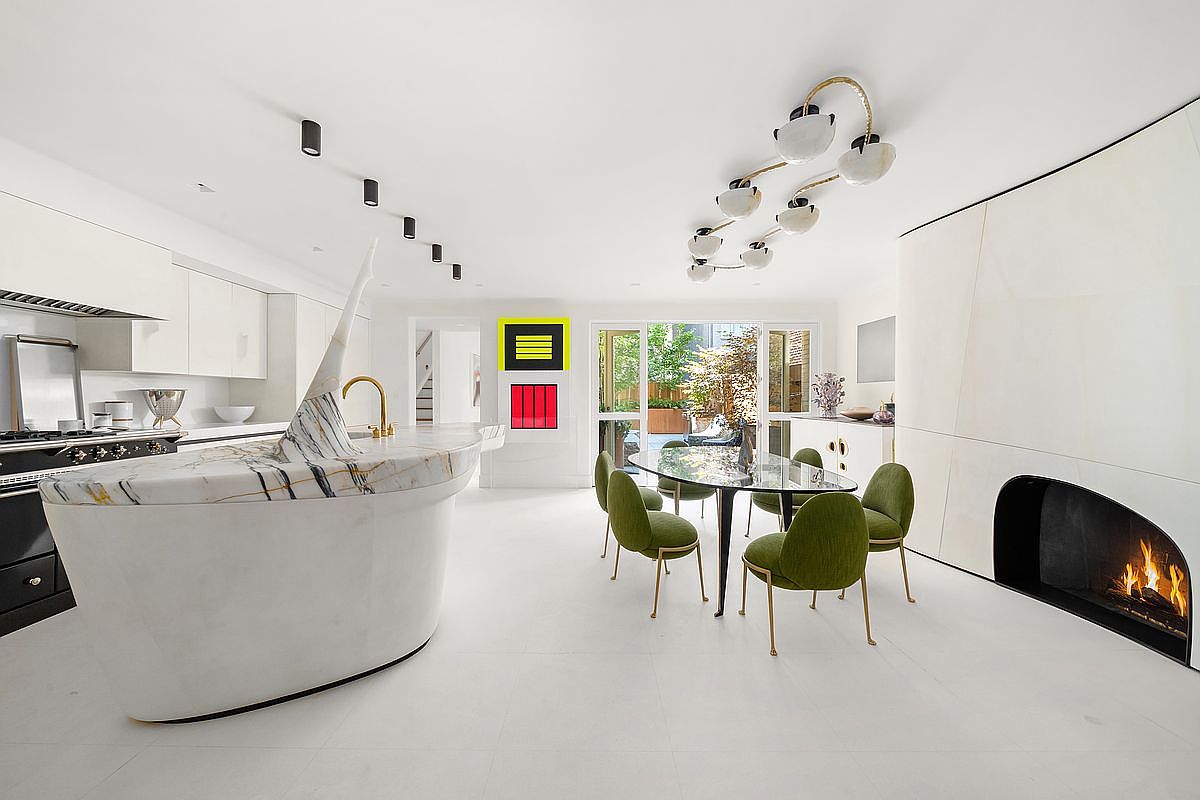 This expansive, open-concept kitchen and dining area features a striking, curved stone-topped island with a dramatic, sculptural accent piece. The space is anchored by a minimalist fireplace with a tall, smooth surround and a glass-topped dining table surrounded by vibrant green chairs. Large sliding glass doors at the rear provide a seamless transition to an outdoor patio, while a unique, multi-globe chandelier adds a modern artistic touch to the bright, white-toned interior.