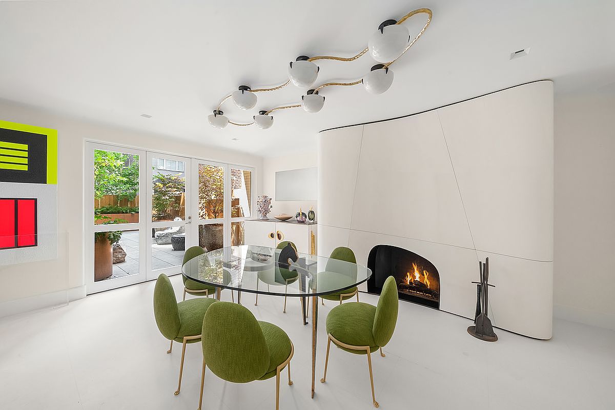 This modern dining room features a glass-topped table surrounded by four vibrant green upholstered chairs, set against a minimalist white backdrop. A striking, sculptural light fixture with multiple globes hangs from the ceiling, while a curved, white wall panel houses a fireplace with a glowing fire. Large glass doors provide a clear view of the outdoor patio, creating a bright and airy atmosphere with a sophisticated, artistic aesthetic.