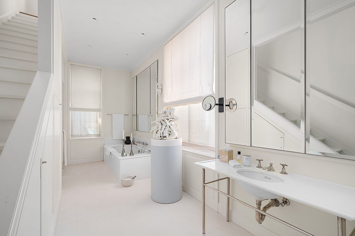 This bright, minimalist bathroom features a sleek white vanity with a pedestal-style sink and a large, multi-panel mirror. A deep soaking tub is positioned near a window, complemented by a unique sculptural pedestal display in the center of the room. The space is characterized by its clean lines, neutral color palette, and a staircase visible on the left, creating an airy and sophisticated atmosphere.