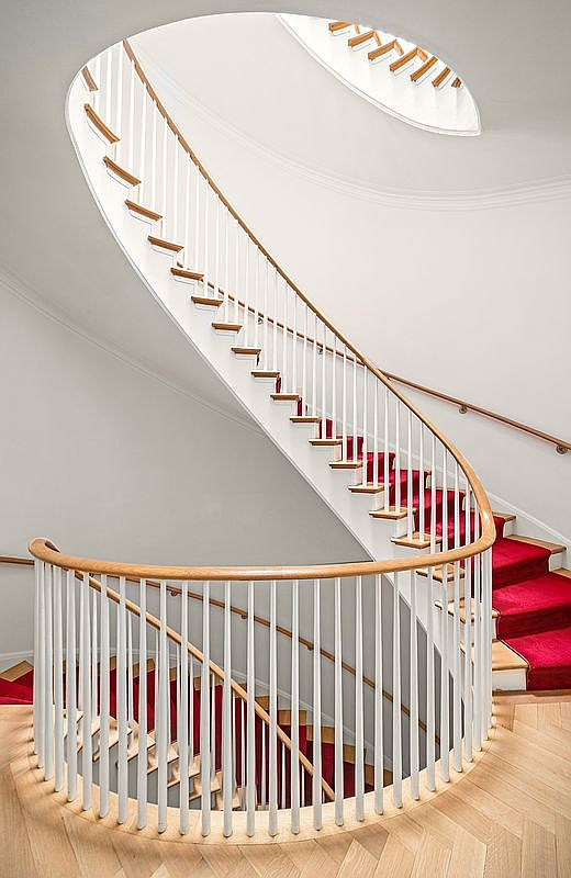 This elegant, curved staircase serves as a dramatic architectural centerpiece, featuring white balusters and a warm-toned wooden handrail. The stairs are lined with a vibrant red runner that contrasts against the light wood flooring and neutral walls. The perspective looks down the spiral, highlighting the graceful, sweeping design and the bright, airy atmosphere created by the overhead light source.