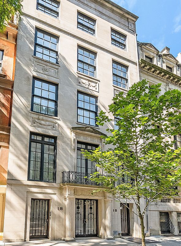 This image captures the elegant front facade of a multi-story townhouse, characterized by its light-colored stone exterior and symmetrical architectural design. The building features tall, multi-pane windows, a decorative balcony with black iron railings on the second level, and a grand entrance framed by classical columns. A mature tree stands in the foreground, partially obscuring the lower right side of the structure, while the perspective is captured from a low angle looking up to emphasize the building's height and sophisticated urban aesthetic.