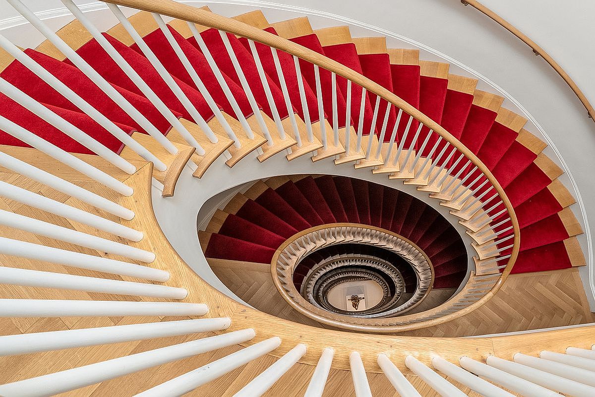 This high-angle, downward-looking shot captures a dramatic spiral staircase featuring light wood treads and a vibrant red runner. The elegant design includes white balusters and a curved wooden handrail that follows the circular geometry of the stairwell. The perspective emphasizes the architectural depth and the rhythmic, repeating pattern of the steps as they descend into the center of the home.