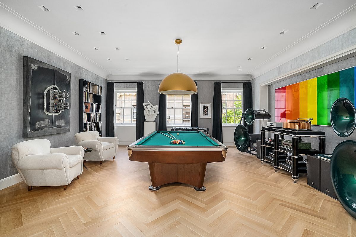 This sophisticated game room features a central pool table illuminated by a large, gold-toned pendant light, set against a backdrop of light wood herringbone flooring. The space is decorated with bold, contemporary art, including a vibrant rainbow-colored panel and a large abstract piece, alongside high-end audio equipment with prominent horn speakers. Two plush white armchairs provide comfortable seating, while the room is framed by dark curtains and tall windows that allow natural light to fill the space.