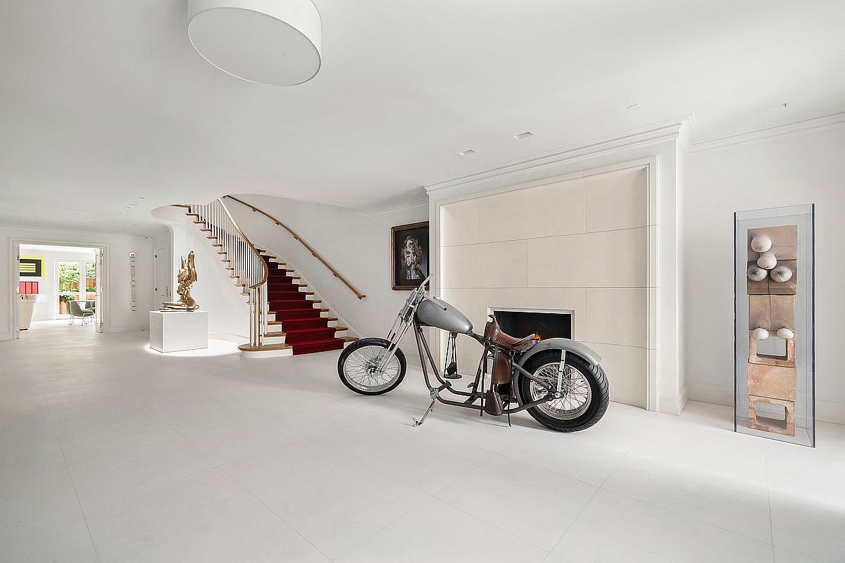 This expansive, minimalist living space features a striking custom motorcycle displayed as a centerpiece in front of a fireplace with a tall, neutral-toned stone surround. A grand staircase with a red runner and a curved wooden handrail sweeps upward, while a sculptural art piece sits on a pedestal nearby. The room is finished with bright white walls and light-colored flooring, creating a gallery-like atmosphere that emphasizes the unique architectural and decorative elements.