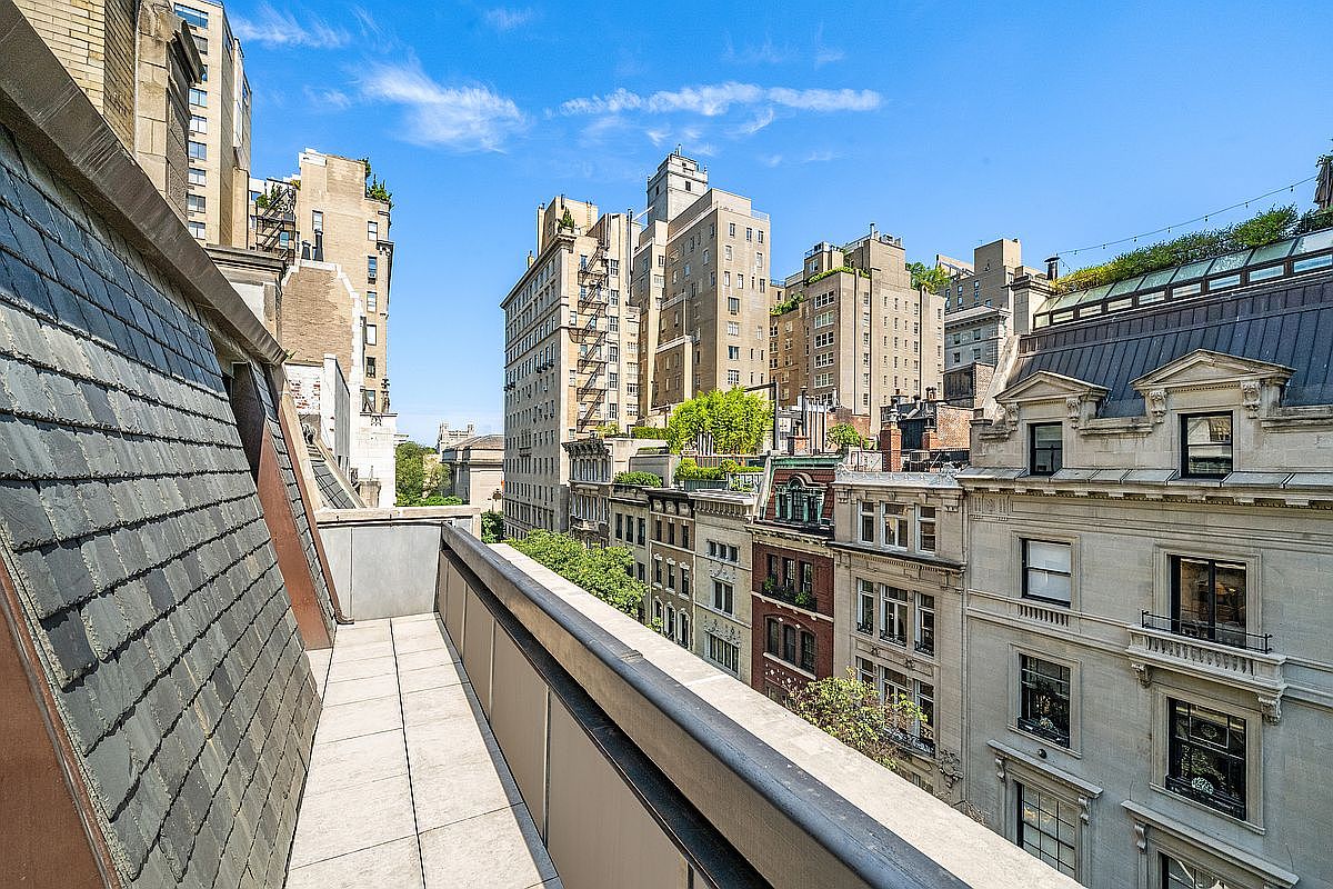 This image captures a private balcony overlooking a dense urban cityscape. The balcony features light-colored stone tiling and a simple metal railing, providing a clear vantage point of the surrounding historic architecture and rooftops. The perspective is elevated, emphasizing the height and the expansive, bright blue sky above the city.