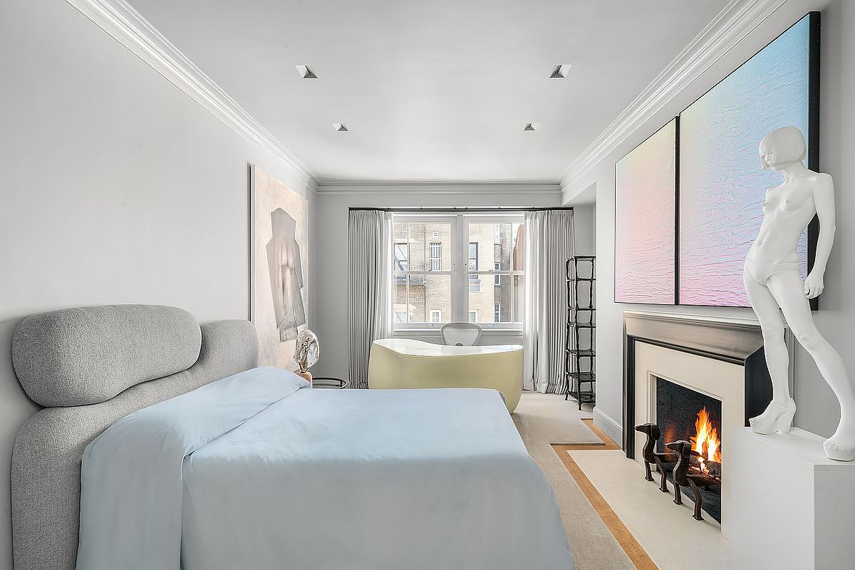 This sophisticated primary bedroom features a plush, curved-back bed with light blue bedding, positioned centrally in the room. A unique, sculptural bathtub sits near the window, while a fireplace with a dark surround and a striking white statue on the hearth adds a dramatic artistic flair. The room is bathed in soft, neutral tones, creating a serene and high-end atmosphere.