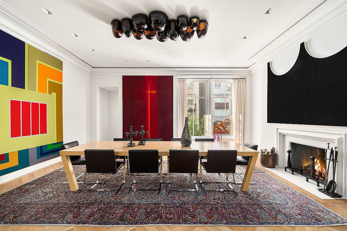 This sophisticated dining room features a large, light wood table surrounded by eight black leather chairs, set atop a detailed, traditional-style patterned rug. The room is anchored by a fireplace with a black surround and a unique, dark, multi-bulb sculptural chandelier hanging above. Bold, contemporary art pieces adorn the walls, creating a striking contrast with the classic architectural details and the view of the street through the window.