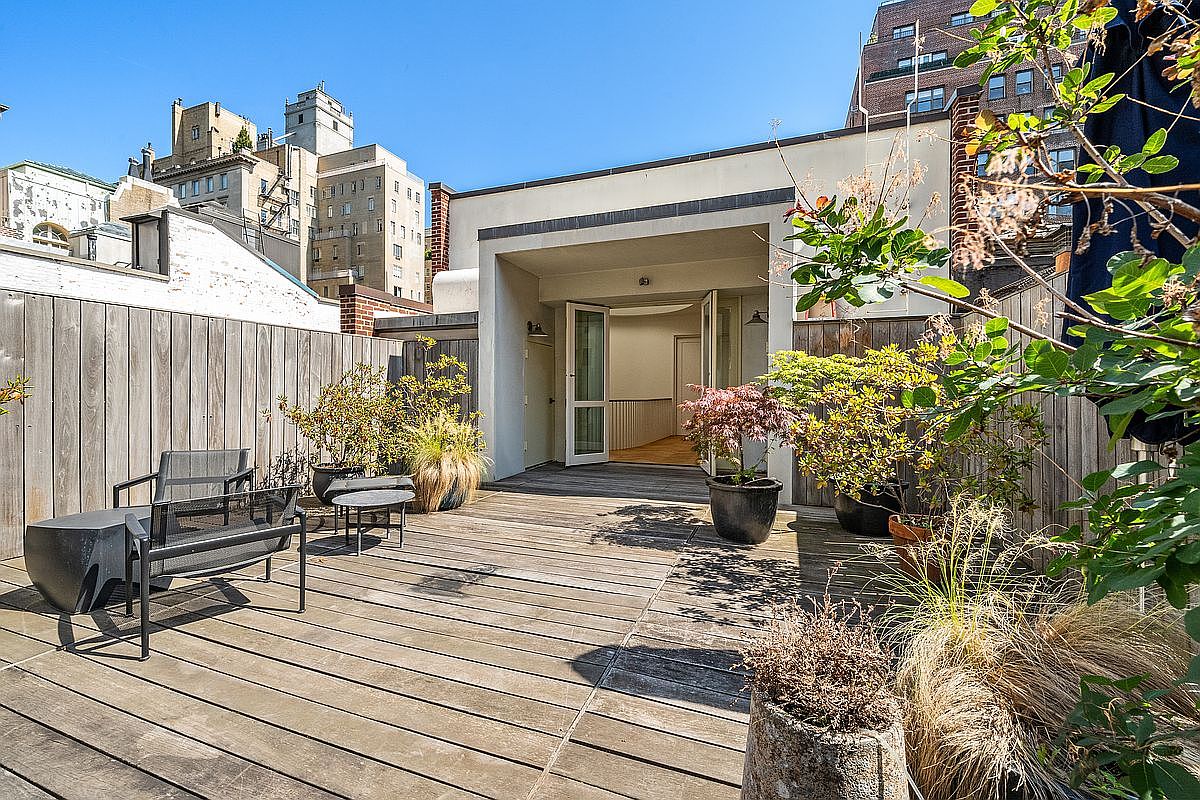 This urban rooftop deck features weathered wood decking and a modern, minimalist aesthetic. The space is furnished with a sleek metal chair and bench set, complemented by various potted plants and small trees that add greenery to the outdoor area. A recessed, covered entryway leads directly into the interior, framed by a clean, light-colored structure that contrasts with the surrounding city skyline.