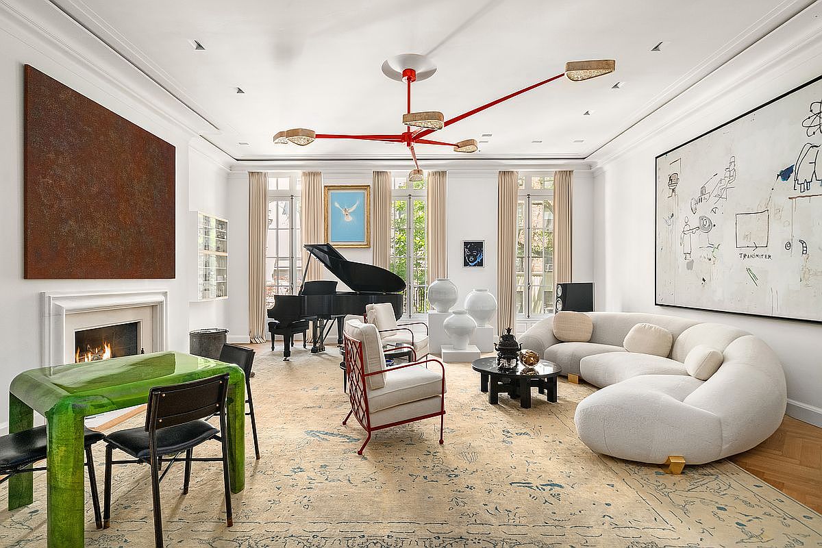 This expansive, high-ceilinged living room features a sophisticated blend of contemporary art and classic architectural details. The space is anchored by a large, curved white sofa, a grand piano, and a striking red multi-armed chandelier, all set against a backdrop of tall windows draped in neutral curtains. A large, textured area rug defines the seating area, which includes a glass-topped dining table and a fireplace with a minimalist surround, creating an atmosphere of refined luxury and artistic flair.
