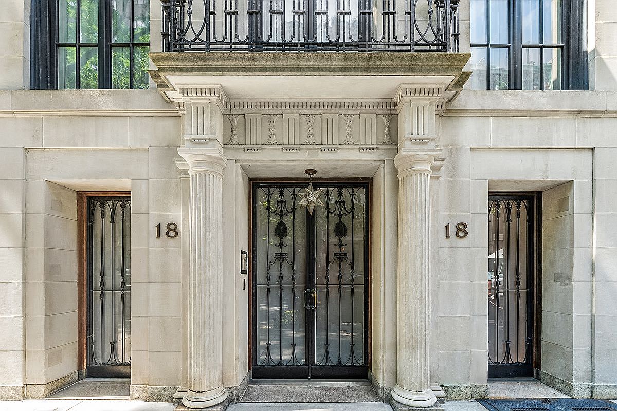 This elegant building entrance features a symmetrical facade with a central double door framed by two classical columns. The entryway is adorned with intricate ironwork grilles on the doors and flanking side windows, while a decorative star-shaped light fixture hangs above the main entrance. The stone exterior, marked with the number 18 on both sides, conveys a sophisticated and historic architectural style.