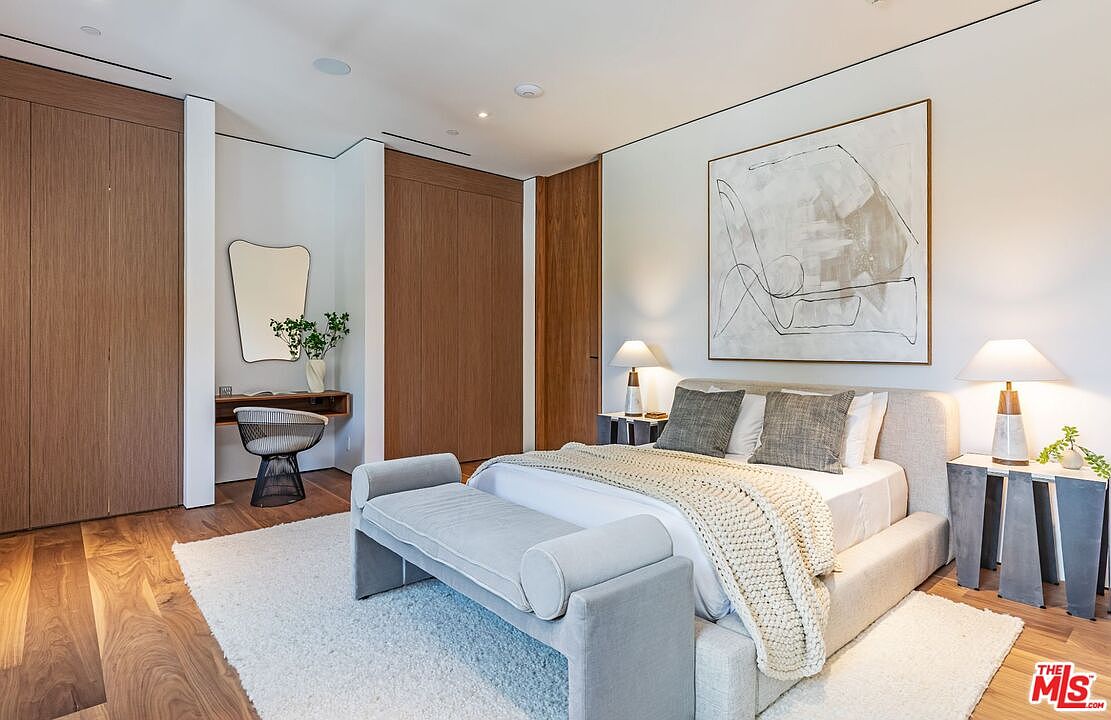 This sophisticated primary bedroom features a neutral color palette, warm wood paneling, and a plush upholstered bed frame. The room is accented by a modern vanity area with a unique mirror, a textured area rug, and minimalist bedside tables with elegant lamps. The overall aesthetic is serene and contemporary, offering a luxurious and tranquil retreat.