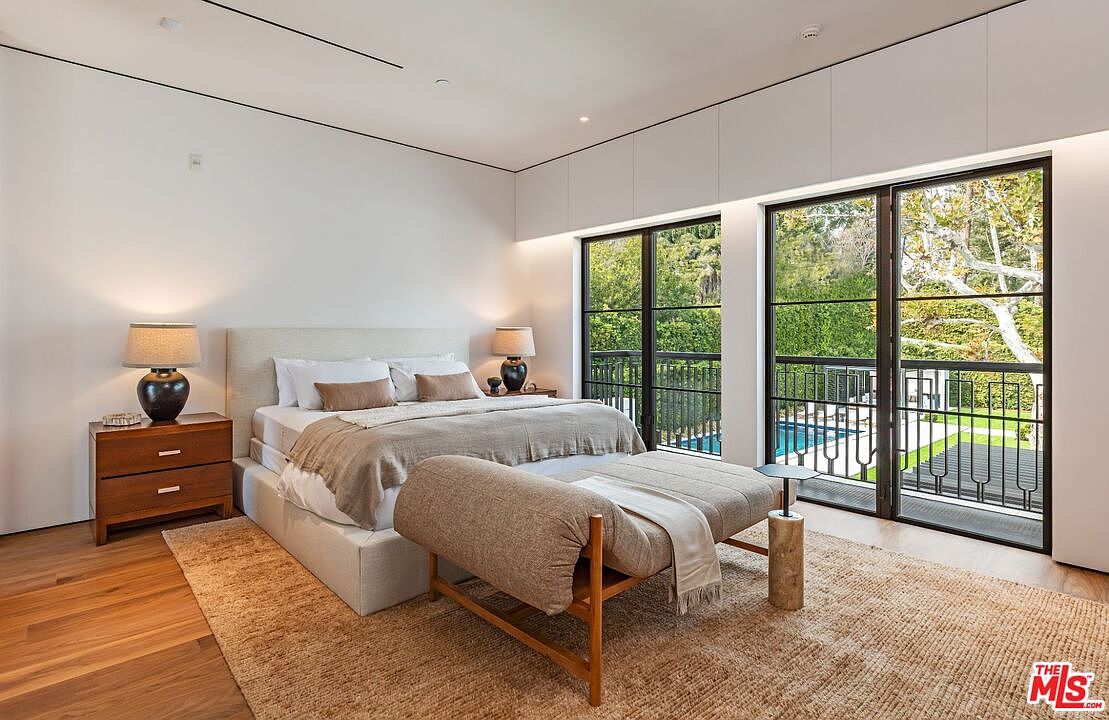 This elegant primary bedroom features a neutral color palette, a plush upholstered bed, and a matching bench at the foot, creating a serene and sophisticated atmosphere. Large floor-to-ceiling glass doors provide abundant natural light and a seamless view of the outdoor pool and lush greenery. The room is anchored by warm wood flooring and a textured area rug, emphasizing a modern, high-end aesthetic.