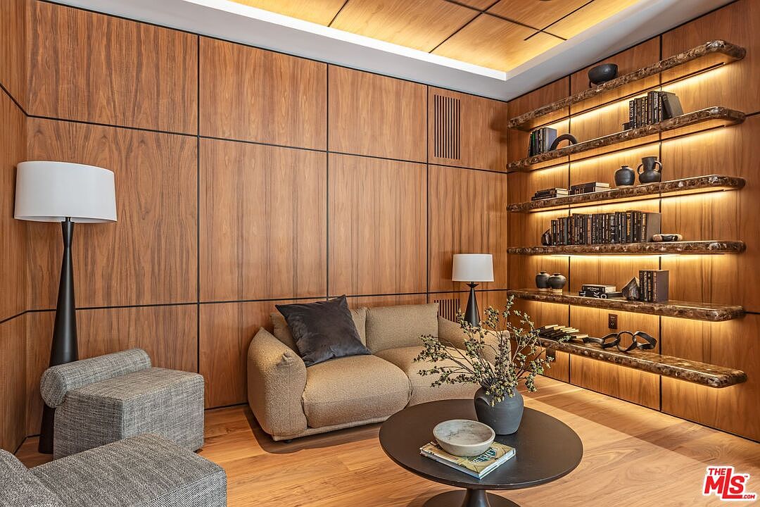 This sophisticated study features warm wood-paneled walls and custom floating shelves with integrated LED lighting, creating a cozy and luxurious atmosphere. The room is furnished with a plush, textured sofa, a modern round coffee table, and a sleek floor lamp, offering a perfect blend of comfort and refined design. The perspective is a wide, eye-level shot that captures the inviting seating area and the elegant display of books and decor.