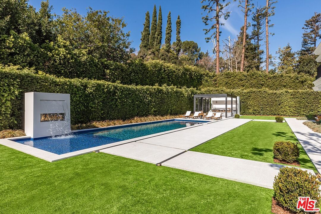 This luxurious backyard features a sleek, rectangular lap pool with a modern water feature integrated into a white architectural wall. A manicured lawn and concrete pathways lead to a contemporary cabana furnished with lounge chairs, all set against a backdrop of tall, lush privacy hedges. The scene exudes a high-end, serene atmosphere perfect for outdoor entertaining and relaxation.