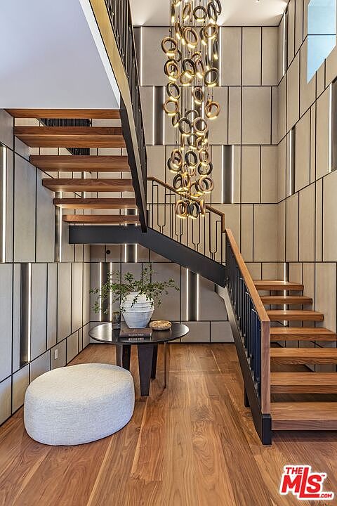 This striking entryway features a dramatic floating staircase with rich wood treads and a sleek black metal railing. A grand, multi-ring chandelier hangs from the high ceiling, casting a warm glow over the space, while a minimalist round table and plush ottoman sit below. The walls are adorned with a sophisticated geometric paneling design accented by integrated vertical lighting, creating a luxurious and modern architectural statement.