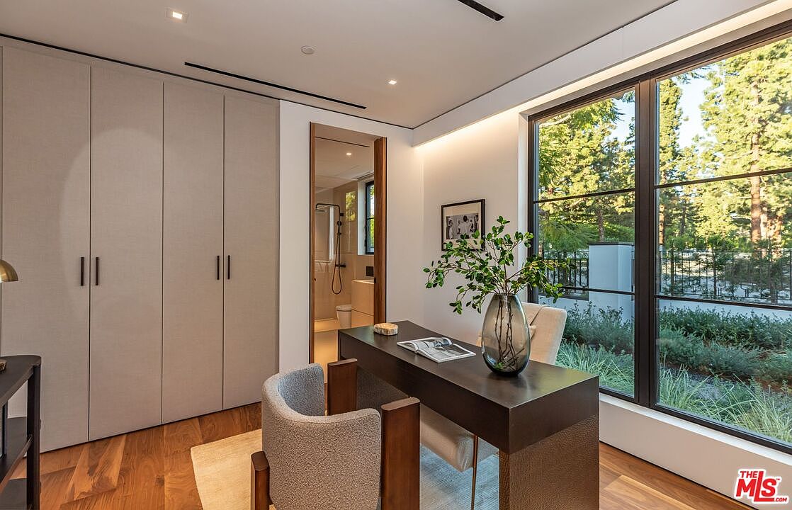 This sophisticated home office features a sleek, dark wood desk positioned to overlook a lush, green garden through expansive floor-to-ceiling windows. The room is accented by a textured, light-colored wall of floor-to-ceiling cabinetry and a curved, upholstered chair, creating a serene and professional atmosphere. The perspective is eye-level, capturing the seamless blend of modern interior design and natural outdoor beauty.