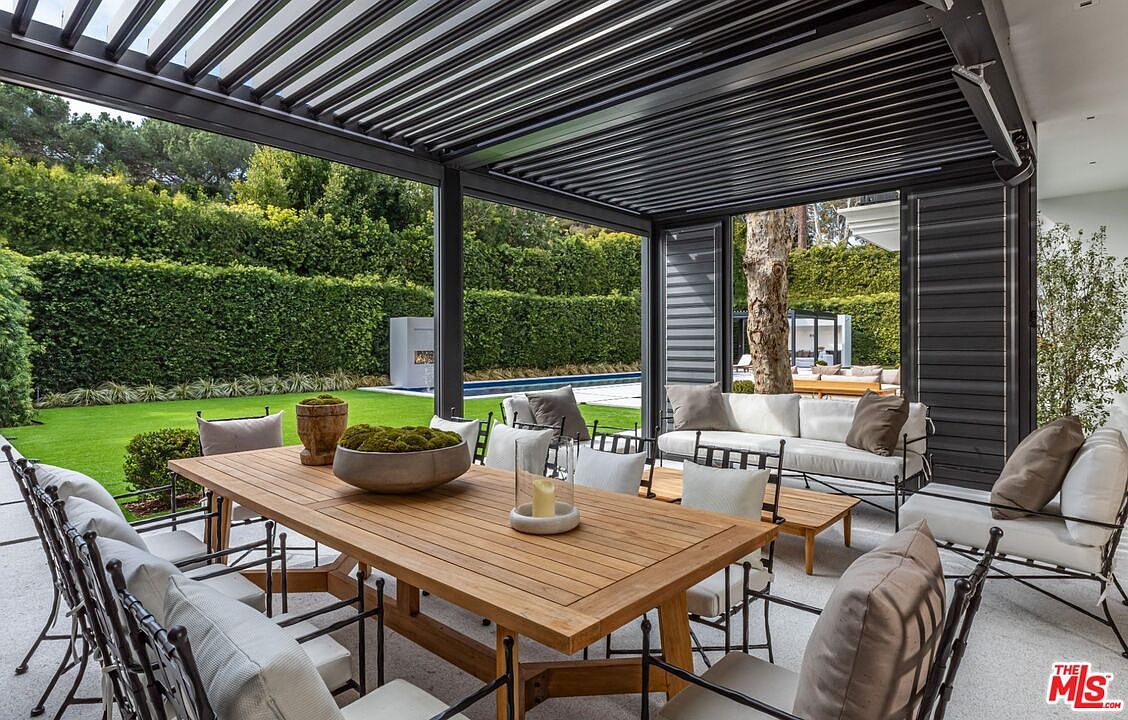 This luxurious covered patio features a sleek, modern louvered pergola roof and a spacious wooden dining table set for outdoor entertaining. The area is furnished with comfortable, neutral-toned lounge seating and overlooks a manicured lawn, a swimming pool, and a lush green hedge, creating a serene and private backyard oasis. The perspective is from the dining area, looking out toward the garden, emphasizing the seamless transition between indoor comfort and outdoor living.