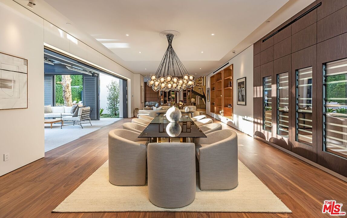 This sophisticated dining room features a long, dark glass-topped table surrounded by plush, light-colored upholstered chairs, all centered beneath a striking, multi-bulb chandelier. The room seamlessly transitions to an outdoor patio through expansive, floor-to-ceiling sliding glass doors, while a wall of dark wood cabinetry with integrated glass-front storage adds a touch of warmth and luxury. The open-concept design and high-end finishes create an elegant, airy atmosphere perfect for formal entertaining.
