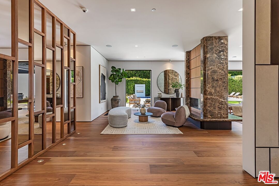 This sophisticated living area features a warm, open-concept design with rich hardwood flooring and a striking marble-clad fireplace column. The space is furnished with contemporary, curved seating and a minimalist coffee table, all arranged on a textured area rug that leads the eye toward a large glass door opening to a lush outdoor patio. A decorative wooden room divider adds architectural interest, while the neutral color palette and soft lighting create an inviting, high-end atmosphere.