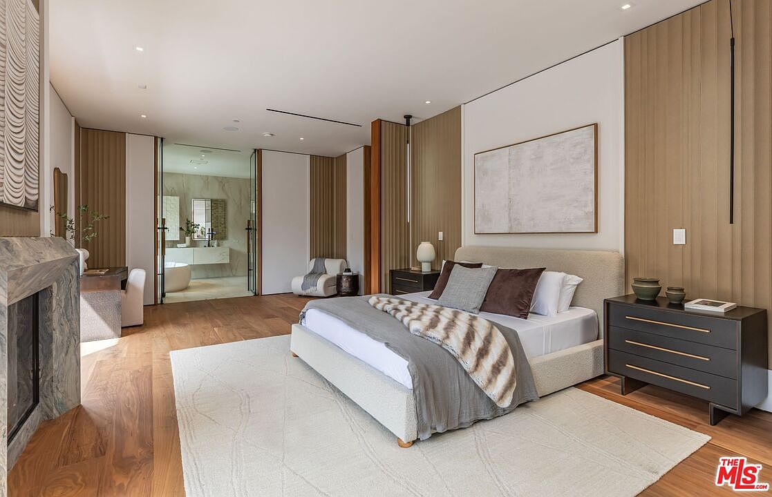 This luxurious primary bedroom features a sophisticated, neutral color palette with warm wood paneling and a textured, upholstered bed frame. A large, plush area rug anchors the space, while a modern fireplace and an open-concept en-suite bathroom with a soaking tub add to the high-end, serene atmosphere. The room is bathed in soft, natural light, creating an inviting and elegant retreat.