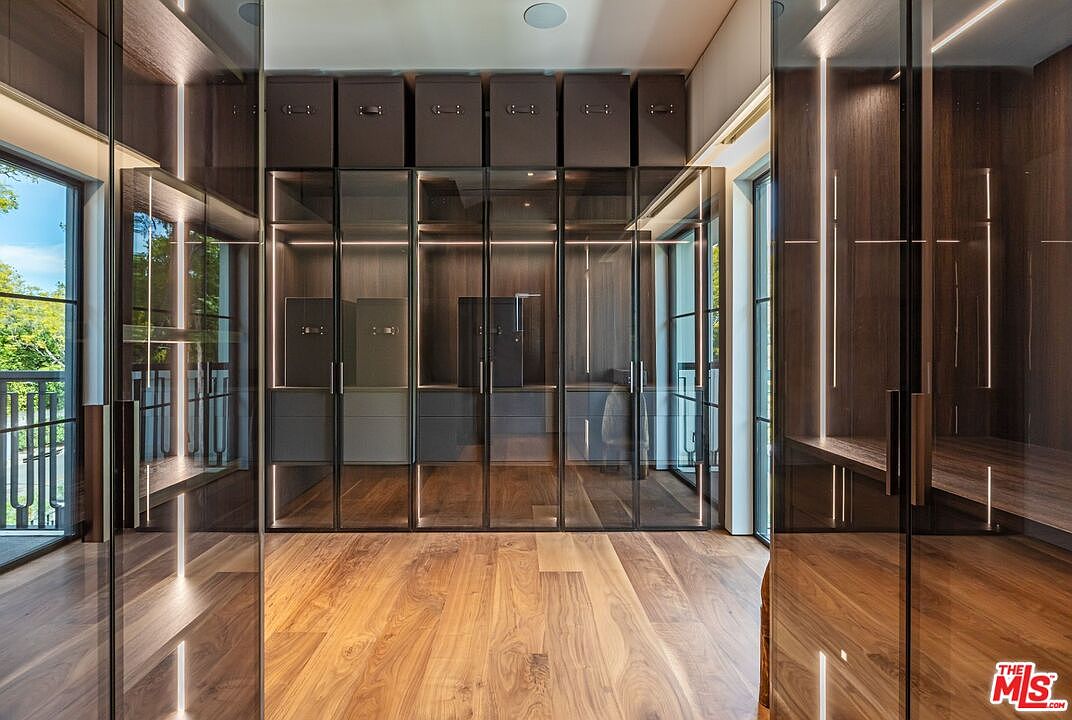 This sophisticated walk-in closet features floor-to-ceiling custom cabinetry with dark wood finishes and sleek glass-front doors, illuminated by integrated vertical LED lighting. The space is anchored by rich, warm-toned hardwood flooring and includes matching storage bins on the top shelf, creating a clean, organized, and high-end aesthetic. The perspective is centered, looking directly into the closet, which is flanked by additional glass-fronted storage units on the sides, emphasizing a luxurious and spacious design.
