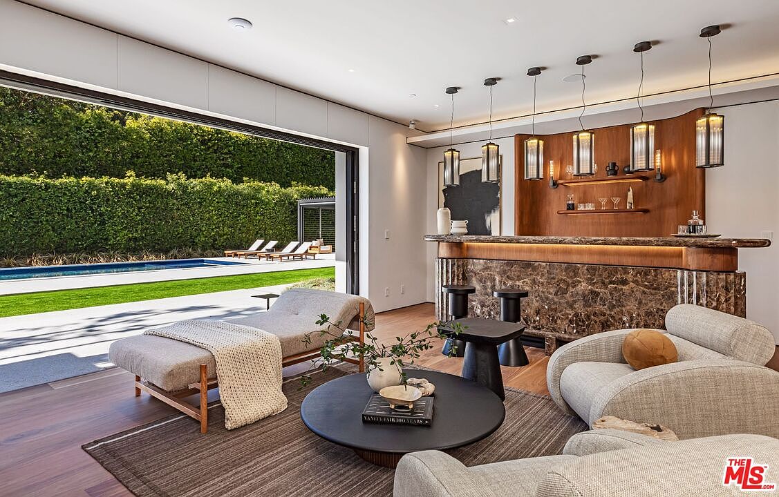 This sophisticated indoor-outdoor living space features a luxurious bar area with marble detailing and warm wood accents, seamlessly transitioning to a sun-drenched patio and pool through expansive sliding glass doors. The interior is furnished with plush, neutral-toned seating and a minimalist coffee table, creating an inviting atmosphere for relaxation or entertaining. The combination of high-end finishes and the direct connection to the lush backyard garden highlights a premium, modern lifestyle.