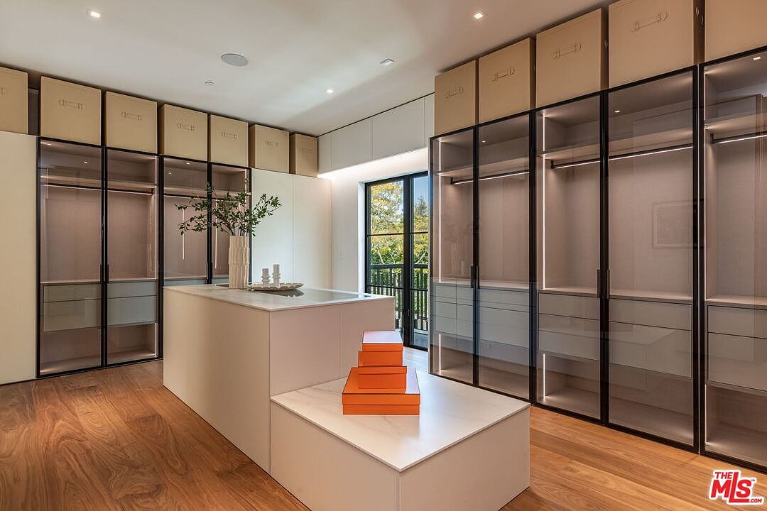 This luxurious, modern walk-in closet features floor-to-ceiling glass-front cabinetry with integrated lighting and a central island topped with white marble. The room is accented by warm wood flooring and a stack of orange designer boxes, creating a sophisticated and organized aesthetic. A large window at the end of the room provides natural light and a view of the greenery outside, enhancing the spacious and high-end feel of the space.