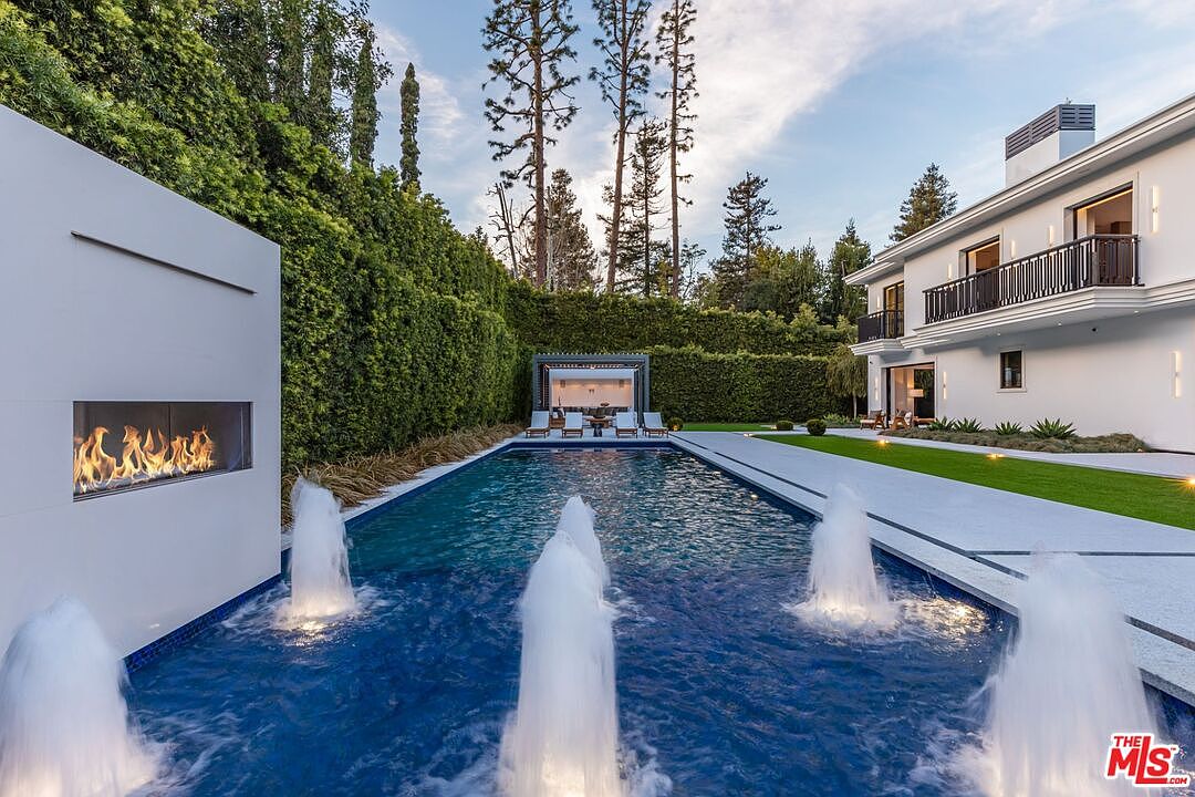 This luxurious backyard features a sleek, rectangular swimming pool accented by multiple water fountains and a modern, built-in outdoor fireplace on the left. The scene is framed by a tall, manicured hedge wall and a contemporary white house with a balcony on the right, creating a private and sophisticated atmosphere. The perspective is a low-angle, eye-level shot that emphasizes the length of the pool and the elegant transition between the water, the lawn, and the architectural elements.