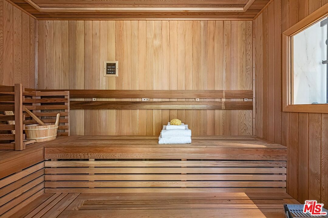This image showcases a traditional cedar-lined sauna, featuring warm wood paneling throughout the walls, ceiling, and tiered benches. A wooden bucket sits on the lower bench, while a neatly folded stack of white towels topped with a natural sponge rests on the upper level, creating a serene and spa-like atmosphere. The perspective is a straight-on, eye-level shot that emphasizes the clean lines and craftsmanship of the wood construction.