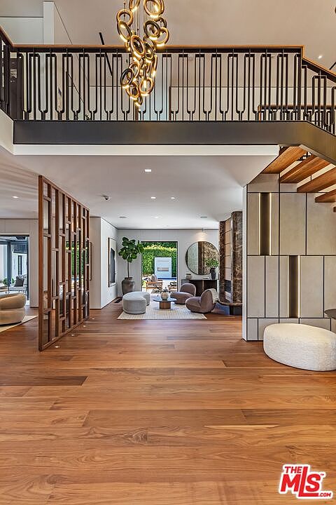 This sophisticated interior view captures a grand entryway and hallway featuring rich hardwood flooring and a striking, modern sculptural chandelier hanging from the upper level. The space is defined by a dramatic open staircase with a sleek black metal railing and a decorative wooden room divider that adds architectural texture. The perspective looks through the open-concept floor plan toward a bright, sunlit living area that seamlessly connects to an outdoor patio, creating an airy and luxurious atmosphere.