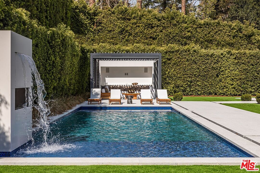 This luxurious outdoor space features a pristine rectangular swimming pool with a modern water feature cascading from a white architectural pillar. Beyond the pool, a sleek, covered cabana provides a shaded lounge area with comfortable seating, all framed by lush, manicured privacy hedges. The scene exudes a high-end, resort-like atmosphere perfect for relaxation and entertaining.