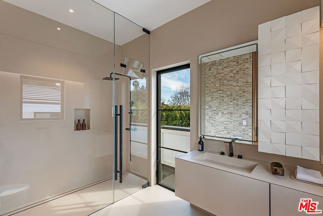 This modern primary bathroom features a sleek, minimalist design with a floating vanity, a large rectangular mirror, and a spacious walk-in glass shower. The room is accented by a textured, geometric tiled wall and a tall, black-framed window that brings in natural light and offers a view of the outdoors. The neutral color palette and clean lines create a sophisticated, spa-like atmosphere.