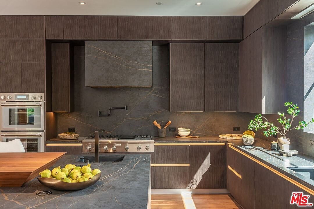 This sophisticated, modern kitchen features dark, wood-grain cabinetry paired with dramatic, dark stone countertops and a matching backsplash with subtle gold veining. A professional-grade stainless steel range and wall oven are integrated into the sleek design, while a large island provides a functional workspace and a bowl of fresh pears adds a touch of organic color. The perspective is a straight-on, eye-level shot that highlights the high-end finishes and the seamless, luxurious aesthetic of the space.