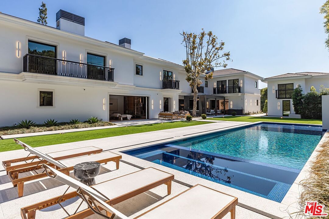 This luxurious backyard features a pristine, rectangular swimming pool with a built-in sun shelf and deep blue tiling, set against a modern white estate. The expansive patio area is furnished with sleek wooden lounge chairs, while a manicured lawn and mature landscaping create a serene, resort-like atmosphere. The architectural design emphasizes clean lines, large glass sliding doors, and multiple balconies, offering a sophisticated and inviting outdoor living space.