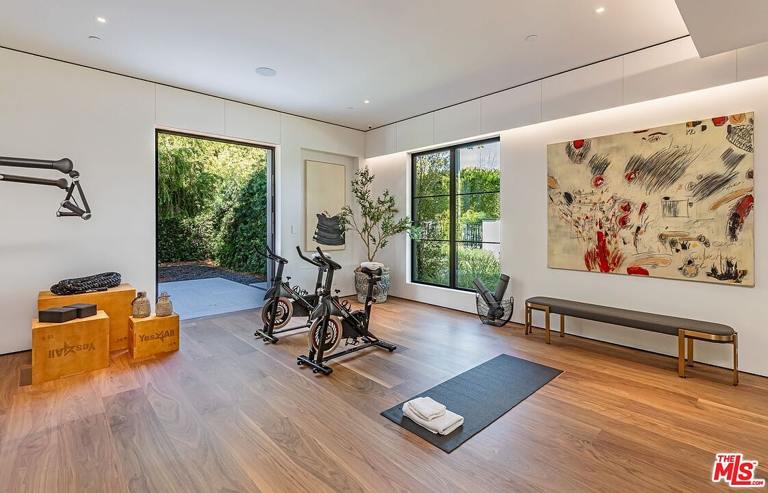 This bright and airy home gym features high-end exercise equipment, including two stationary bikes and wooden plyometric boxes, set against a backdrop of warm hardwood flooring. A large, abstract painting adds a vibrant artistic touch to the minimalist white walls, while a wide doorway opens to a lush outdoor garden, creating a seamless indoor-outdoor workout experience. The space is well-lit by natural light from both the large window and the open door, offering a serene and motivating atmosphere.