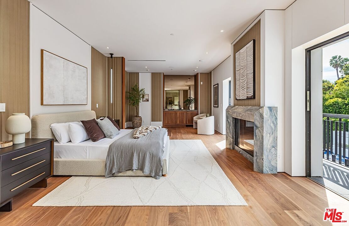This luxurious primary bedroom features a sophisticated, neutral color palette with warm wood paneling and hardwood floors. The room is anchored by a plush bed with textured linens, a striking marble fireplace, and a seamless transition to a private balcony through large glass doors. The space exudes a serene, high-end atmosphere, enhanced by minimalist decor and soft, natural lighting.