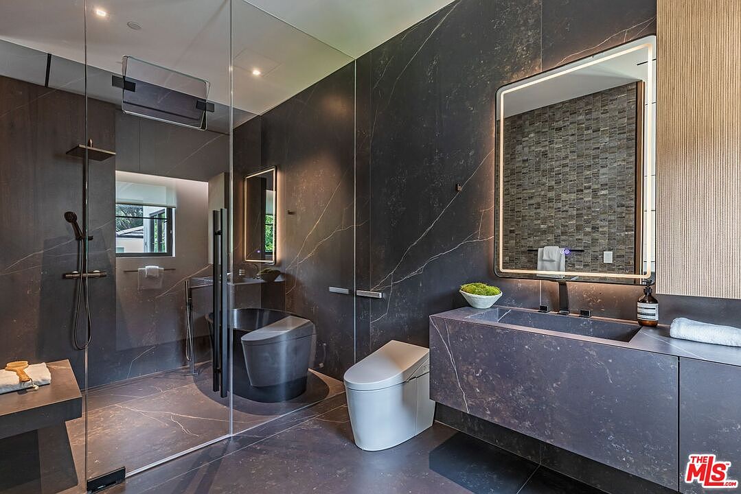 This ultra-modern primary bathroom features dark, veined marble walls and flooring, creating a sophisticated and moody aesthetic. The space includes a sleek floating vanity with an integrated sink, a minimalist wall-mounted toilet, and a glass-enclosed shower area that houses a freestanding soaking tub. A backlit rectangular mirror adds a touch of warmth and luxury to the high-end, contemporary design.