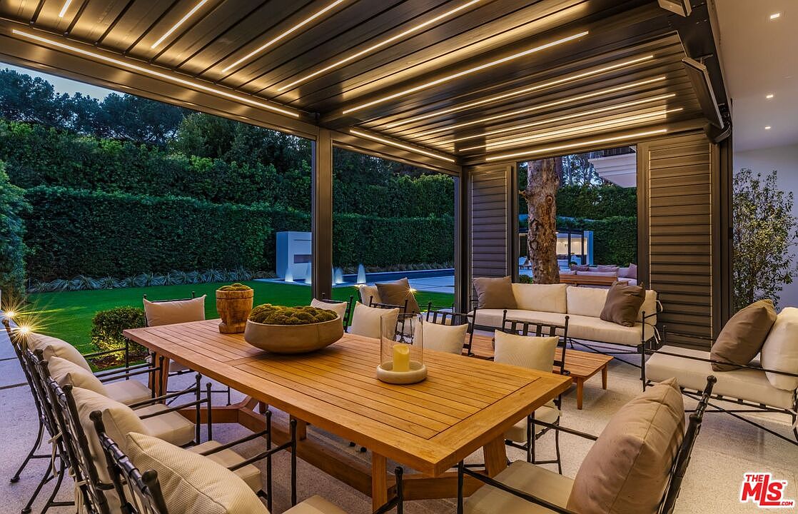 This luxurious covered outdoor patio features a sleek, modern pergola with integrated linear LED lighting, creating an inviting atmosphere for dining and relaxation. The space is furnished with a large wooden dining table and comfortable lounge seating, all overlooking a manicured lawn and a serene pool area. The design seamlessly blends indoor comfort with outdoor living, offering a sophisticated and tranquil retreat.