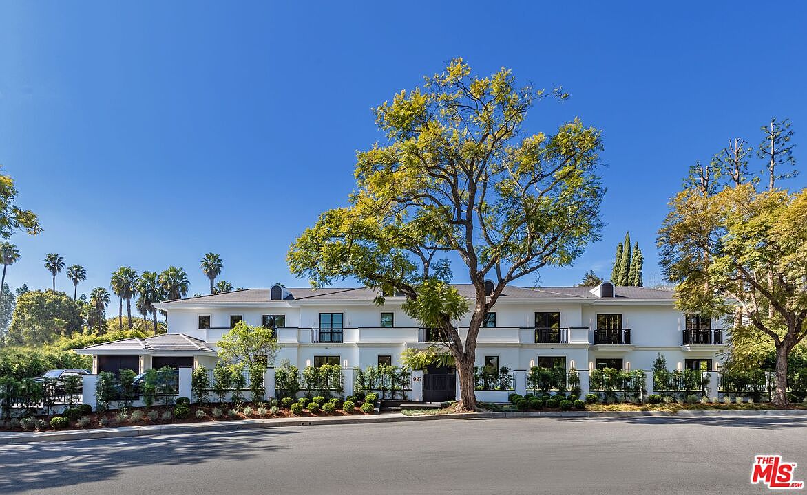 This elegant, two-story white estate features a symmetrical architectural design with a prominent central tree framing the facade. The property includes a gated entrance, manicured landscaping, and a spacious driveway, all set against a clear blue sky. The overall impression is one of luxury, privacy, and sophisticated curb appeal.