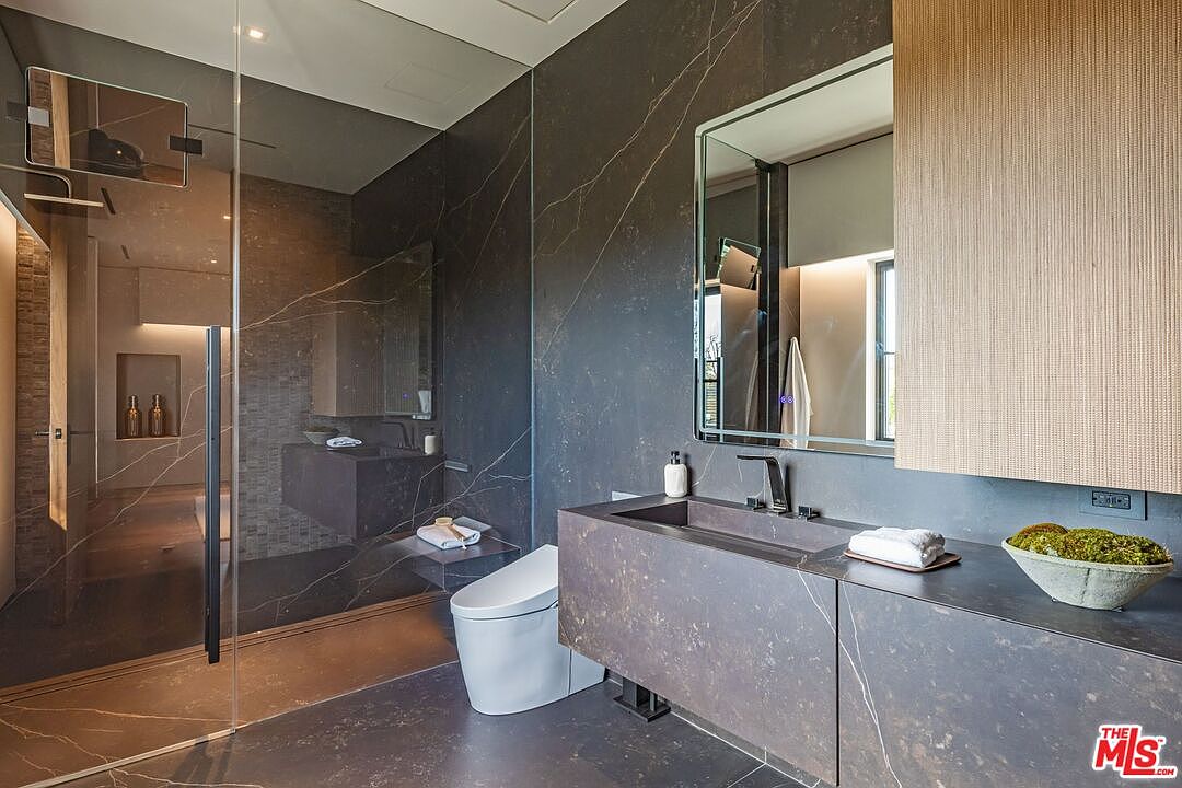 This luxurious, modern bathroom features dark, veined marble walls and flooring that create a sophisticated, moody atmosphere. The space includes a floating vanity with a sleek integrated sink, a minimalist white toilet, and a spacious glass-enclosed walk-in shower. A large, rectangular mirror with integrated lighting and a warm wood-toned cabinet add texture and warmth to the high-end, contemporary design.