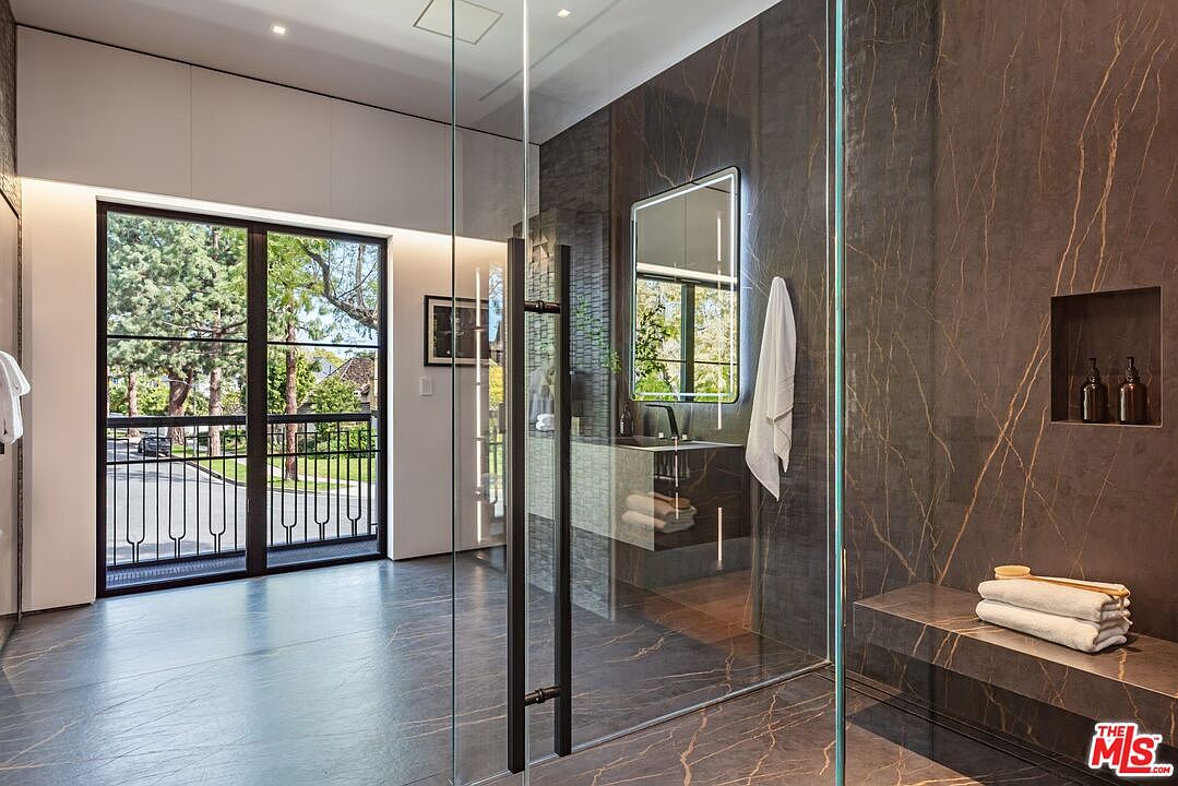 This luxurious primary bathroom features a sleek, modern design with dark, gold-veined marble walls and flooring. A large glass-enclosed shower area includes a built-in stone bench and a recessed niche for toiletries, while a contemporary vanity with an illuminated mirror adds to the sophisticated aesthetic. The space is brightened by a large floor-to-ceiling window that offers a view of the lush greenery outside, creating a seamless blend of indoor elegance and natural beauty.