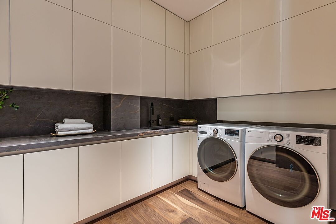 This sleek, modern laundry room features minimalist white cabinetry, a dark stone countertop with an integrated sink, and high-end LG front-loading washer and dryer units. The space is accented by a rich, dark wood-look floor and a sophisticated dark stone backsplash, creating a clean and luxurious aesthetic. The perspective is a wide-angle shot that highlights the efficient layout and high-quality finishes of this functional utility space.