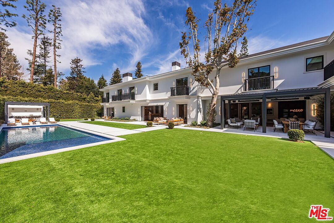 This expansive rear view of a luxury estate showcases a pristine, manicured lawn leading to a modern swimming pool and a covered outdoor patio area. The two-story white residence features clean architectural lines, black-framed windows, and multiple balconies, creating a sophisticated and inviting atmosphere. The scene is captured from a low-angle perspective, emphasizing the seamless transition between the indoor living spaces and the lush, private backyard.