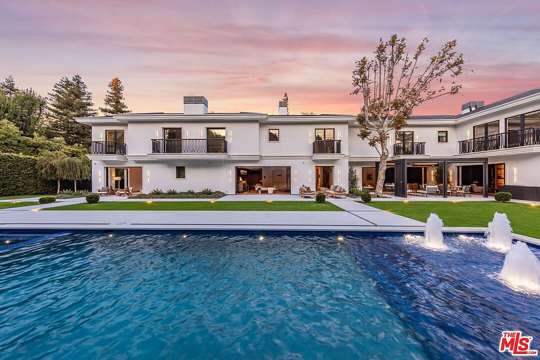 This stunning rear view of a luxury estate showcases a sprawling, modern white mansion set against a vibrant sunset sky. The foreground features a large, pristine swimming pool with active water fountains, leading to a manicured lawn and a seamless indoor-outdoor living space with open glass doors. The architectural design emphasizes clean lines, symmetrical balconies, and a sophisticated, high-end aesthetic perfect for entertaining.